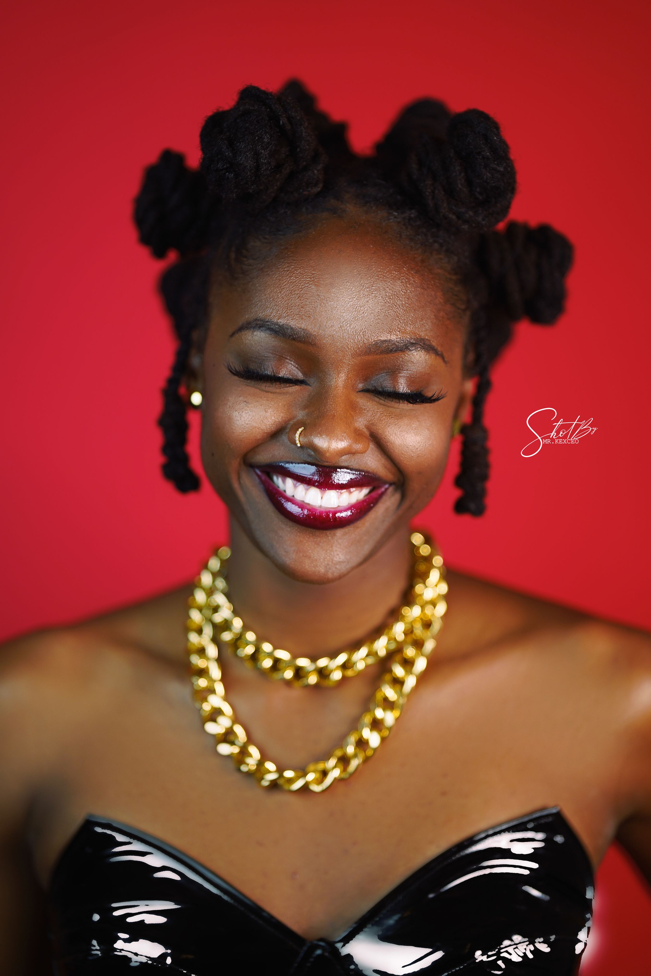Artistic portrait with black hair buns, bold red lipstick and gold chain necklace against red background.