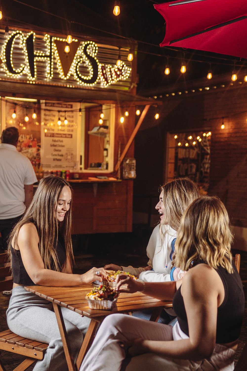 Three friends laugh and share nachos at a cozy outdoor eatery with string lights and a bright sign.