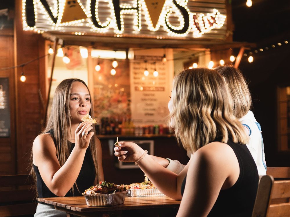 Two young women enjoy nachos together at a warmly lit outdoor restaurant with a glowing NACHOS sign.