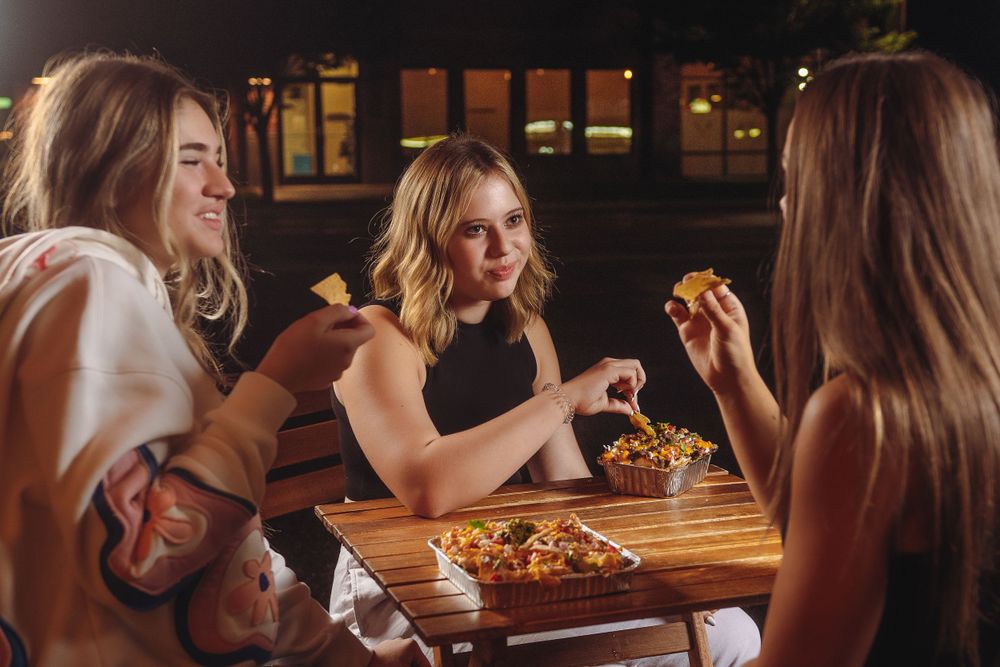 Three young women laugh and snack on nachos together at an outdoor table under night skies.