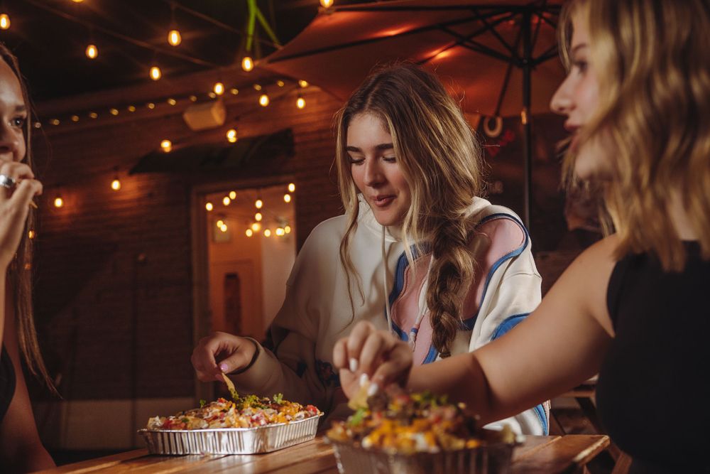 Young women share loaded nachos at an outdoor eatery decorated with warm string lights at night.