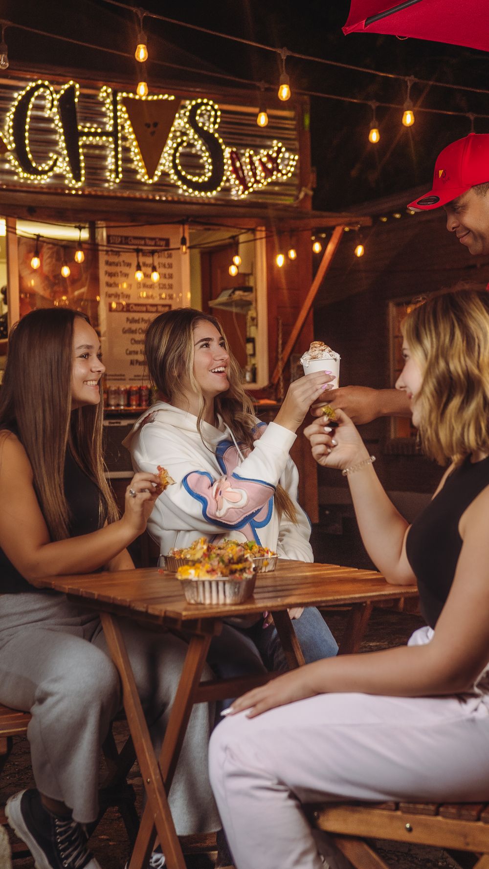 Four friends laugh and share nachos and a milkshake at a glowing outdoor nacho restaurant at night.
