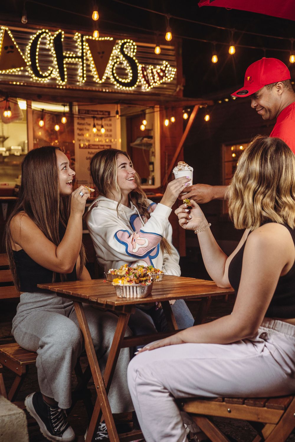Friends laughing and sharing milkshakes at an outdoor restaurant with warm string lights at night.