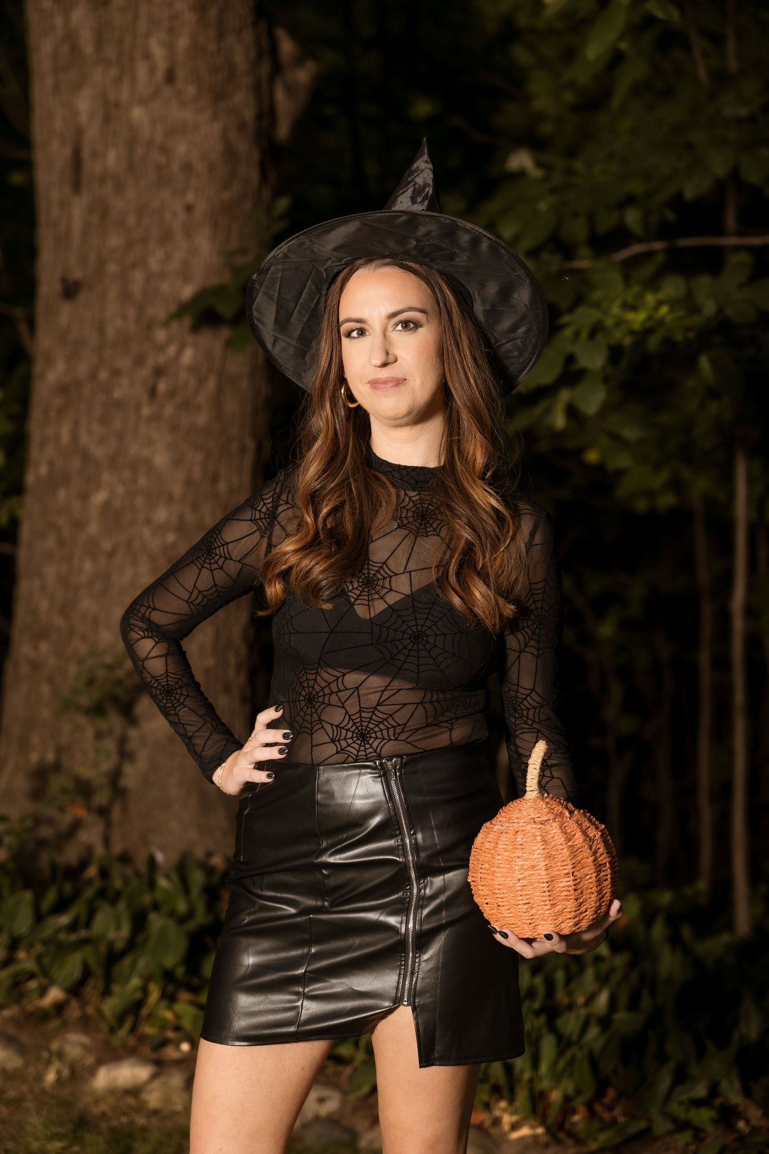 Person in witch hat and leather skirt poses with orange pumpkin prop in outdoor setting.