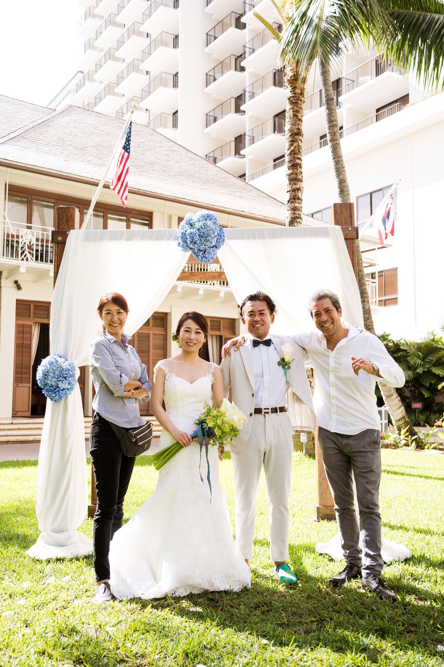 Wedding couple posing with guests under a white floral arch at an outdoor hotel ceremony.