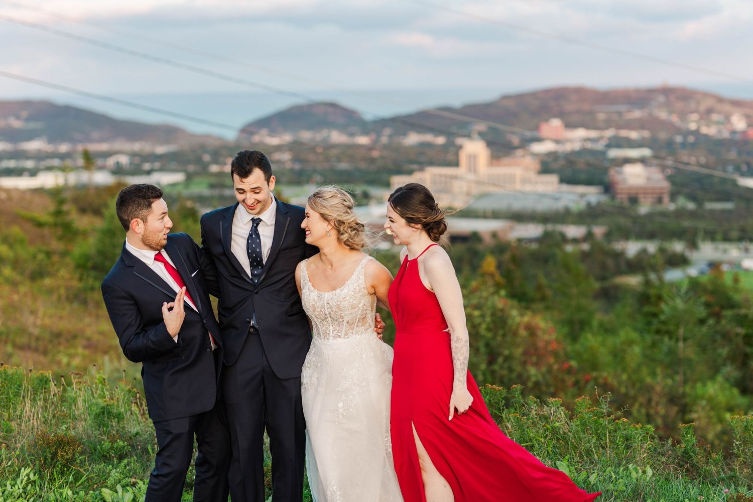 Wedding party members in red and formal attire pose together on a hillside with mountains in the background.