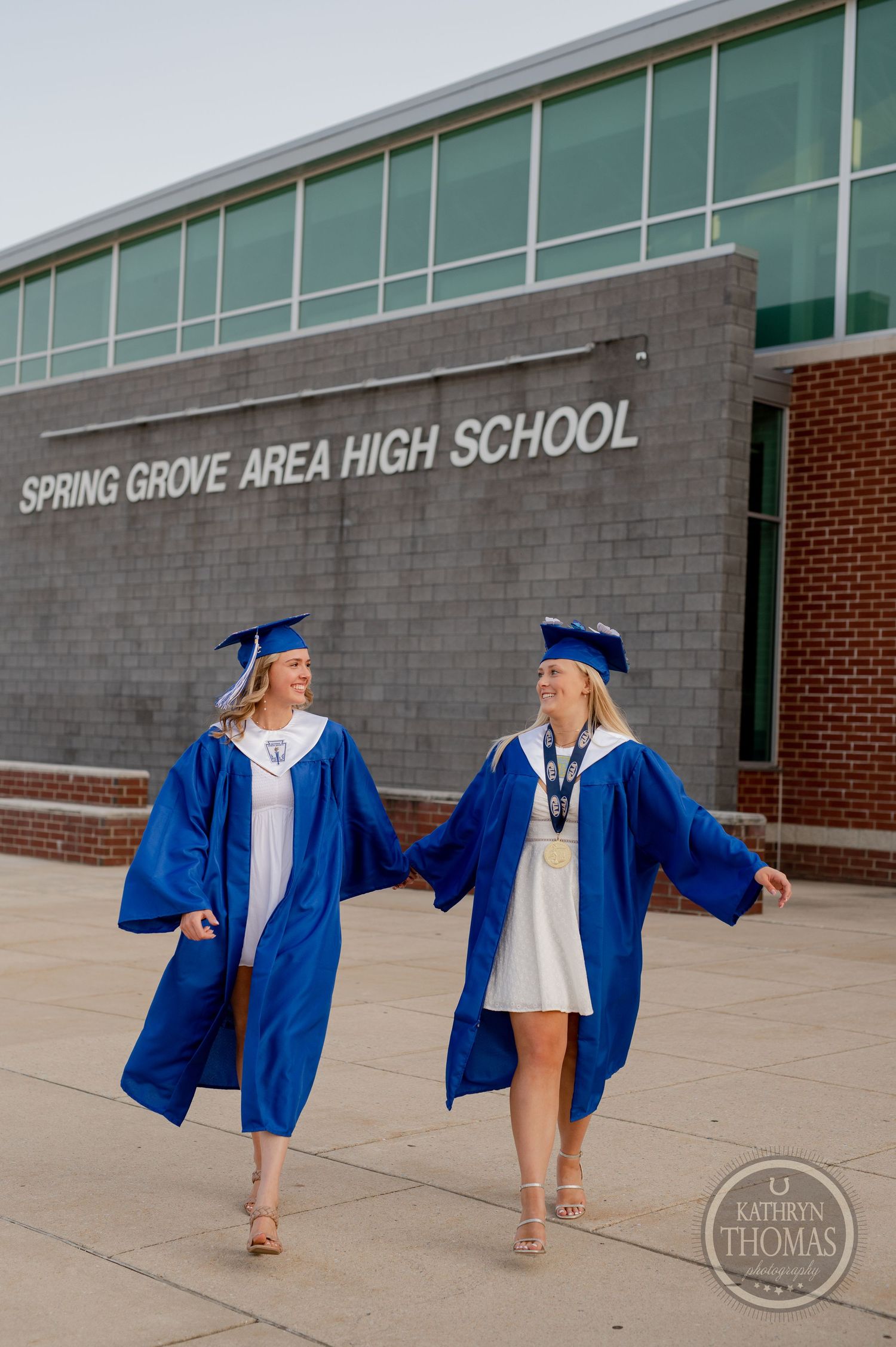 Two graduates in blue regalia hold hands while walking outside their high school.