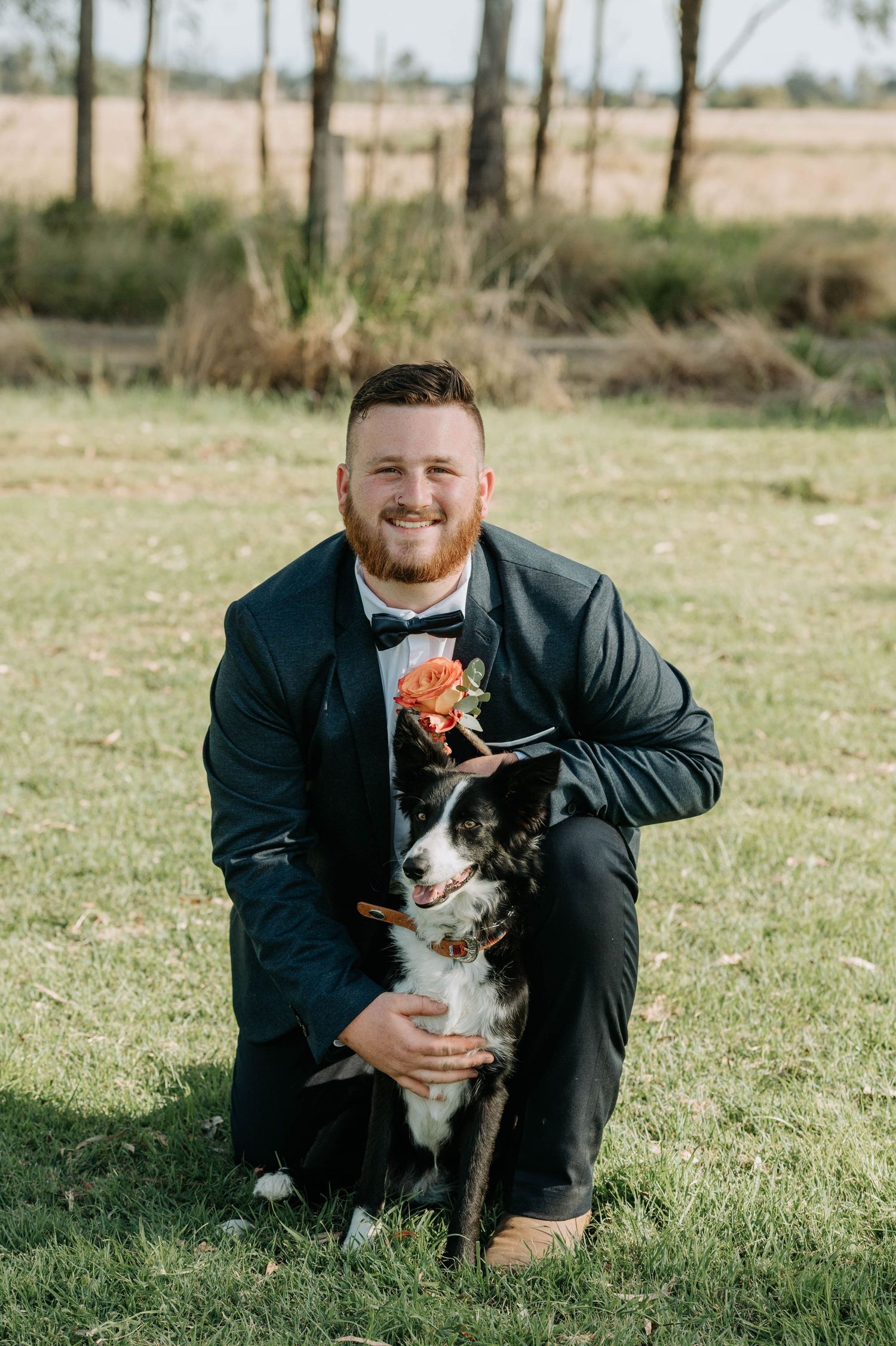 A person in a dark suit kneels with a black and white border collie on grass.