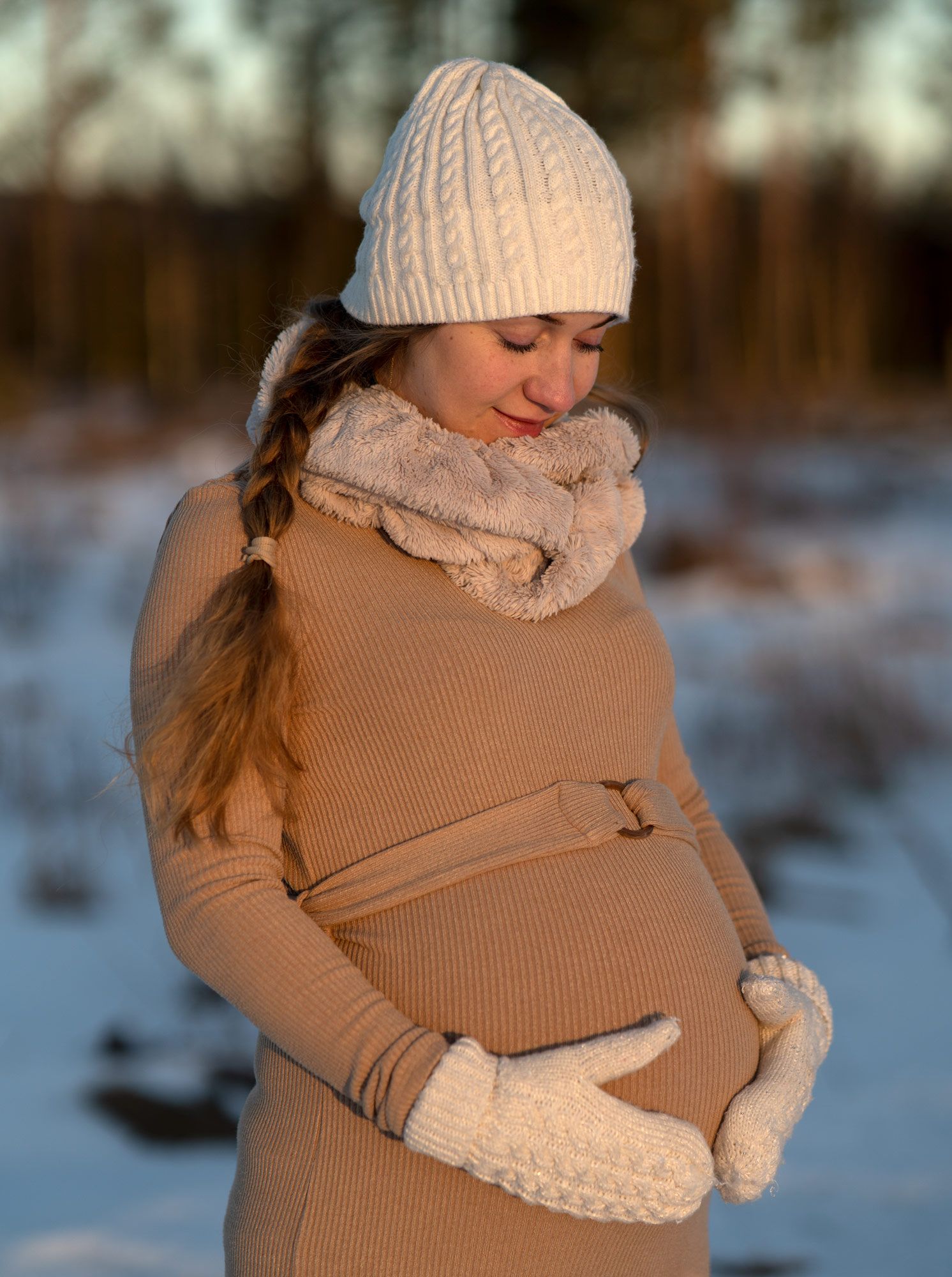 Gravid kvinna i vit mössa och vita vantar håller om sin mage en vinterdag