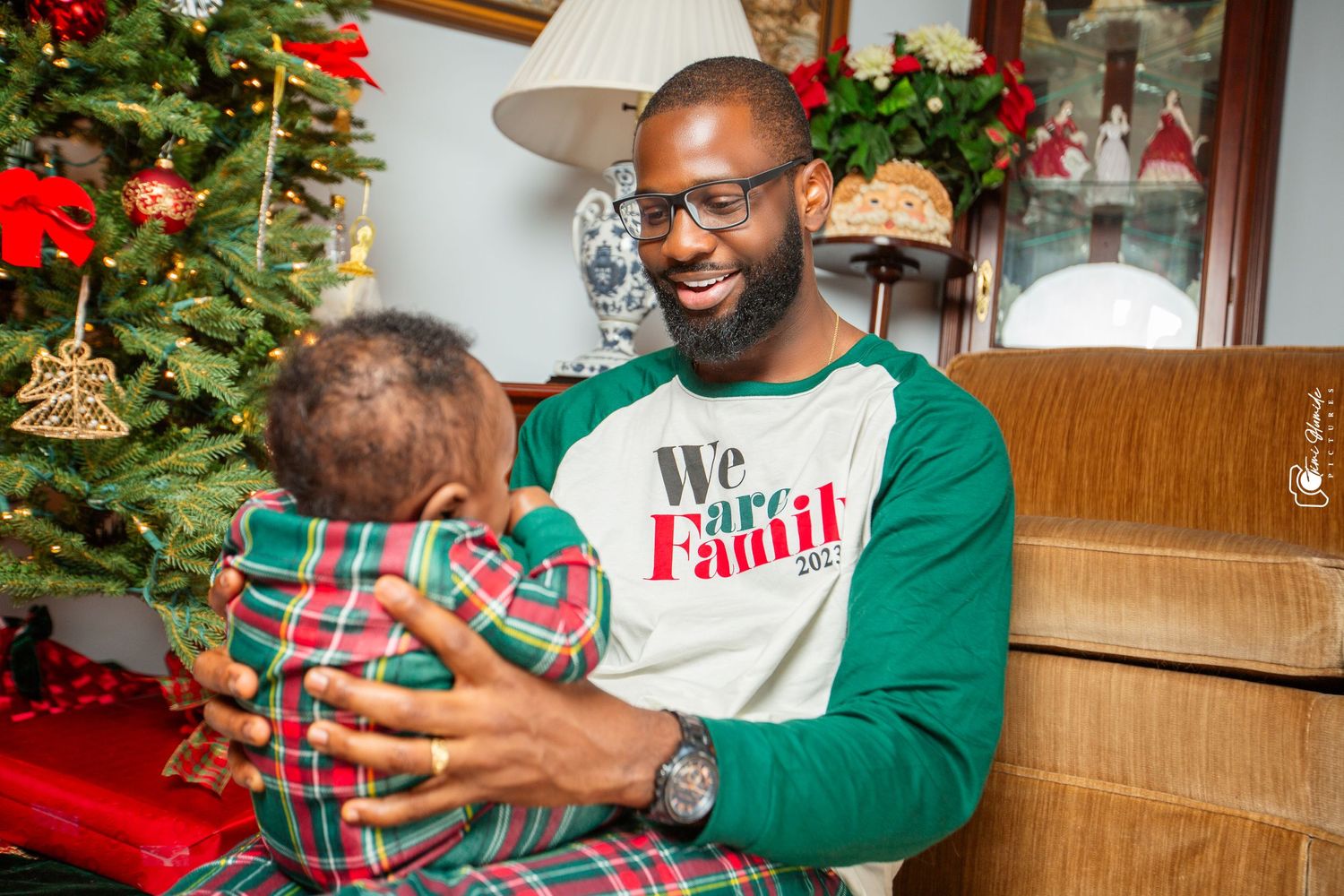 Parent and child in festive clothing share moment by decorated Christmas tree.