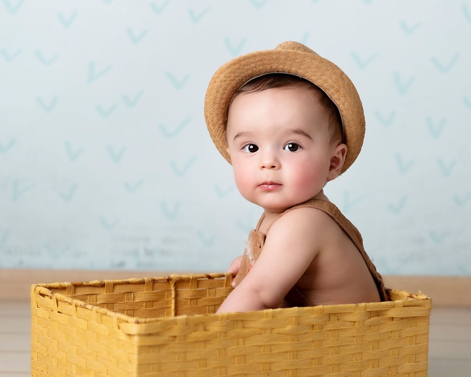 Baby wearing a straw hat sits in yellow woven basket during portrait session.