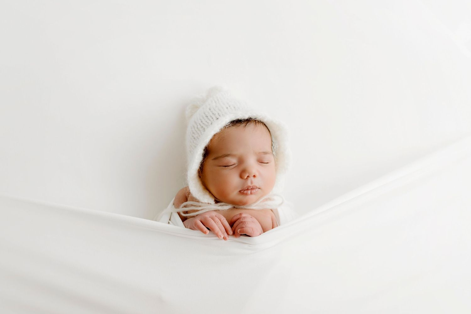 Newborn sleeping peacefully wrapped in white fabric against light background.