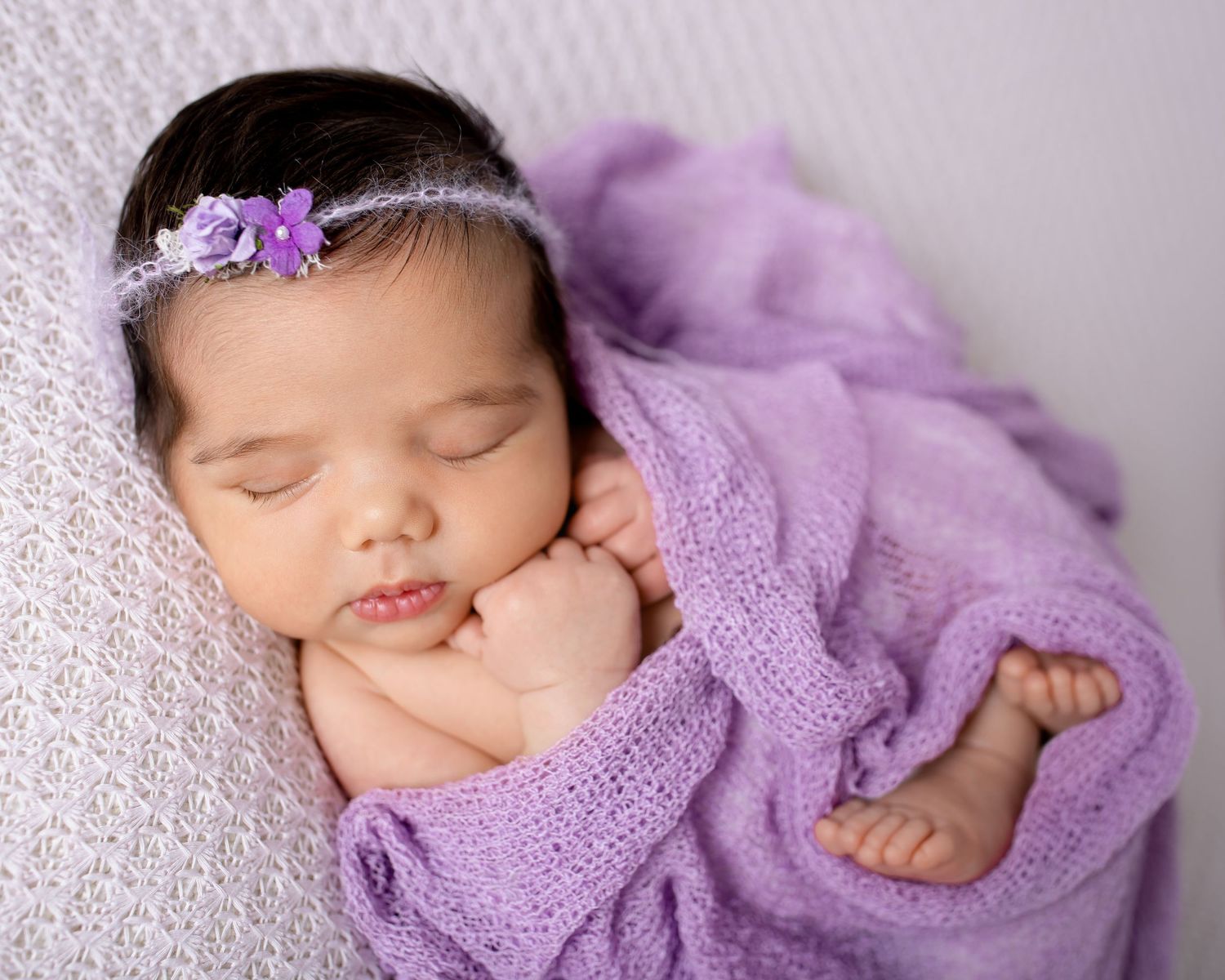 Sleeping newborn wrapped in purple knit blanket with matching hair bow.