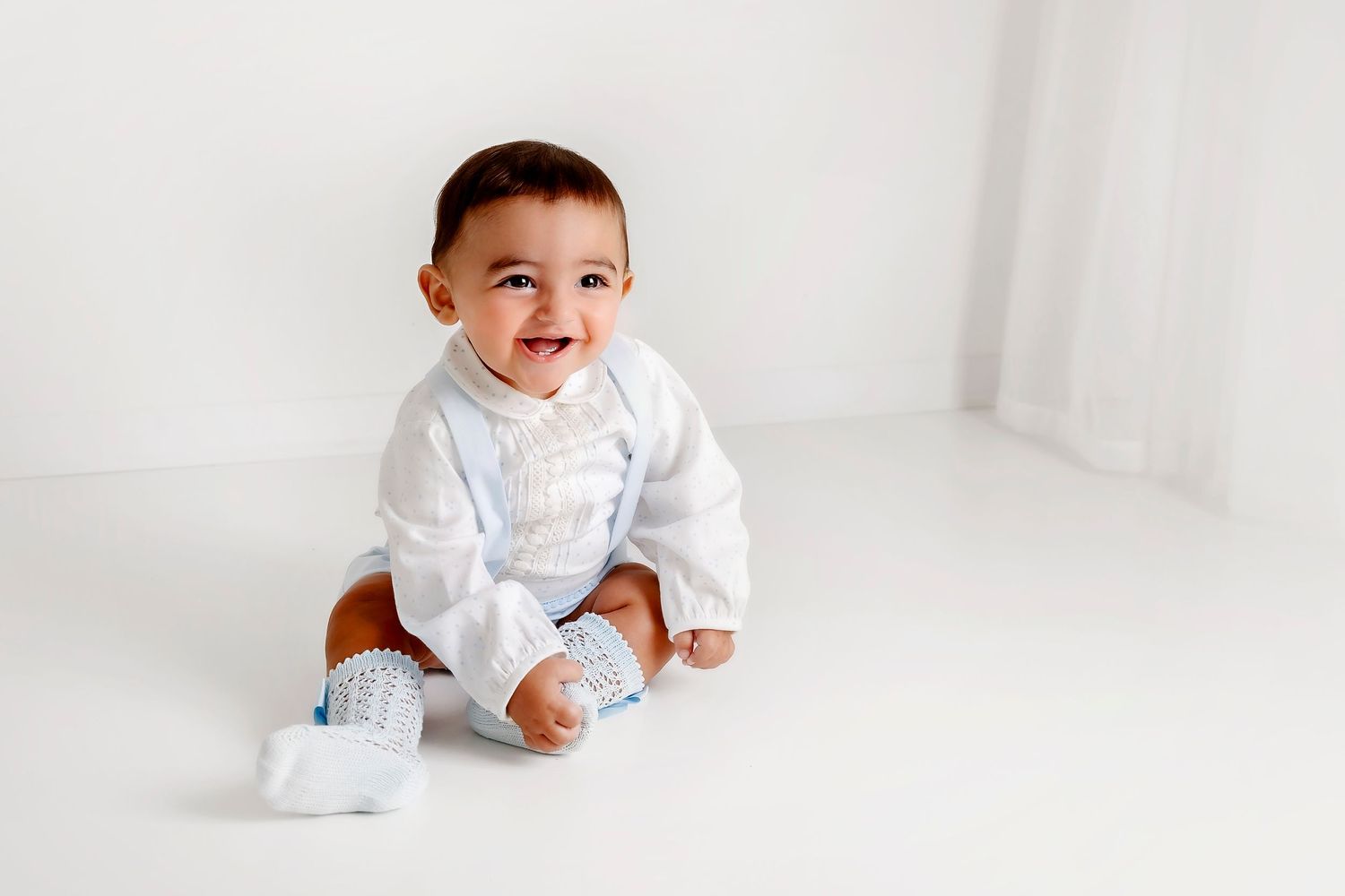 Baby in white outfit sitting against white background during studio photoshoot.