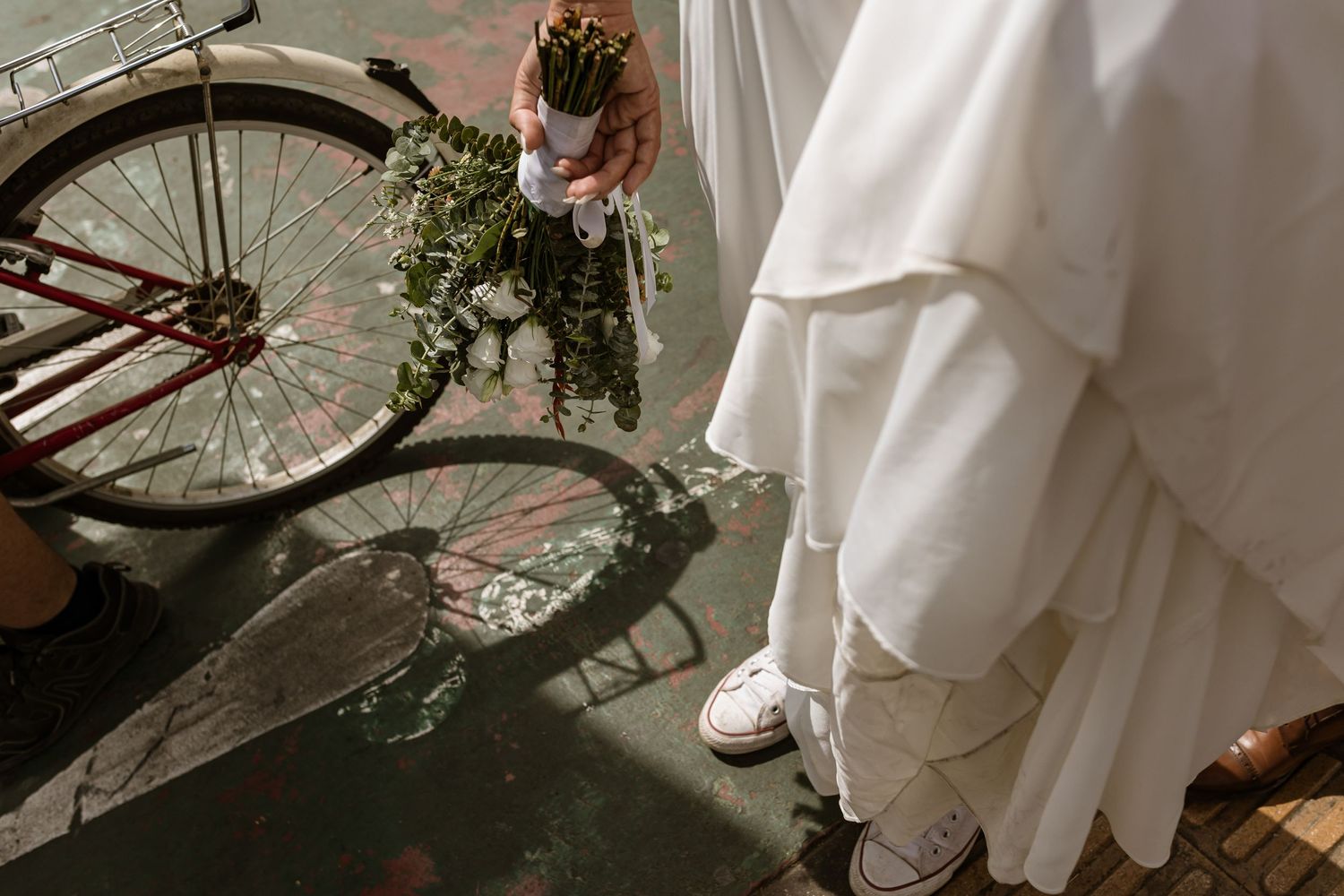 Vintage bicycle and flowing white dress captured in warm sunlight on textured ground with bouquet details.