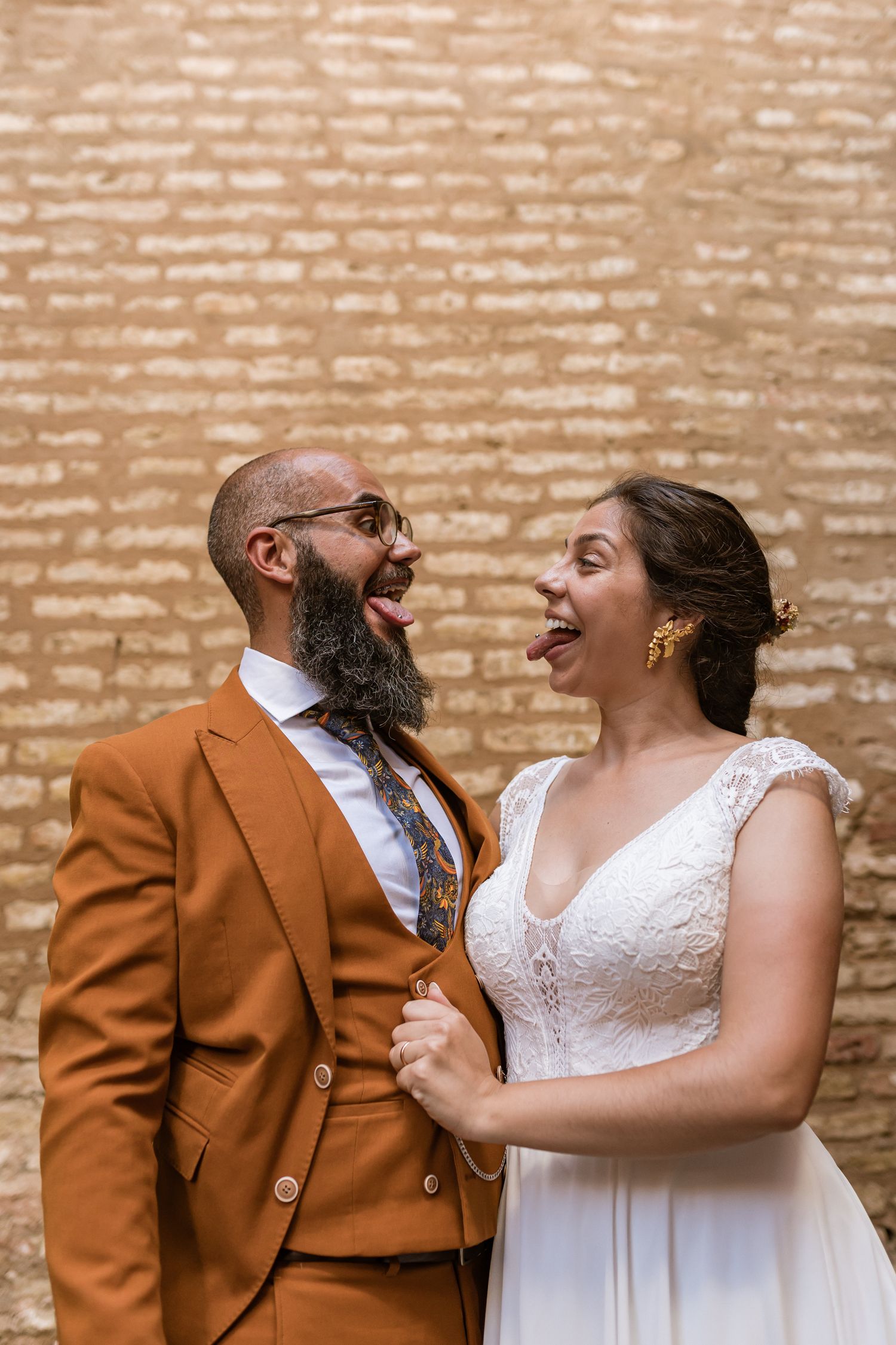 A joyful couple in wedding attire share a laugh against a brick wall background in natural lighting in Seville, Spain