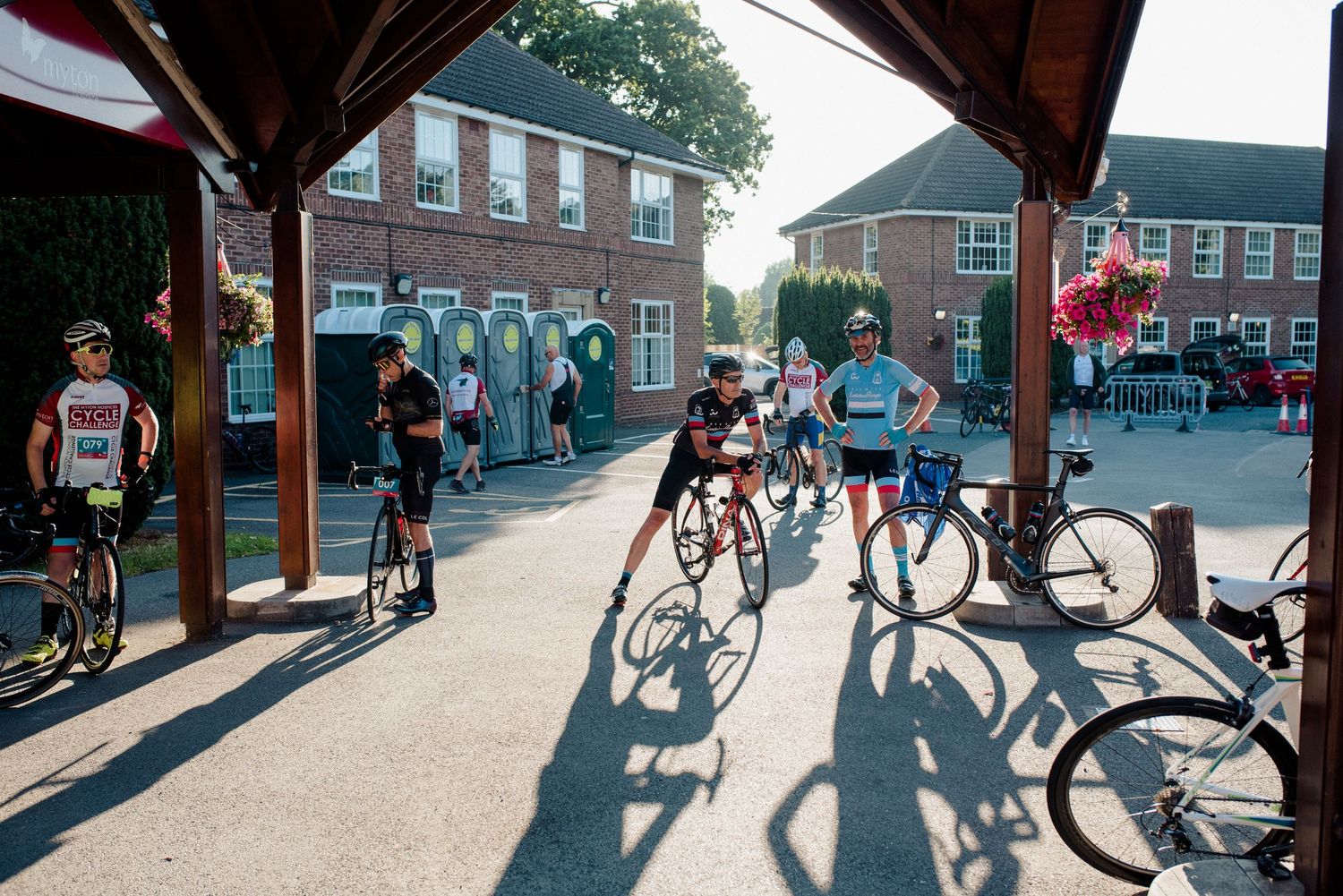 Cyclists gather under a wooden pavilion casting long shadows during a community event.