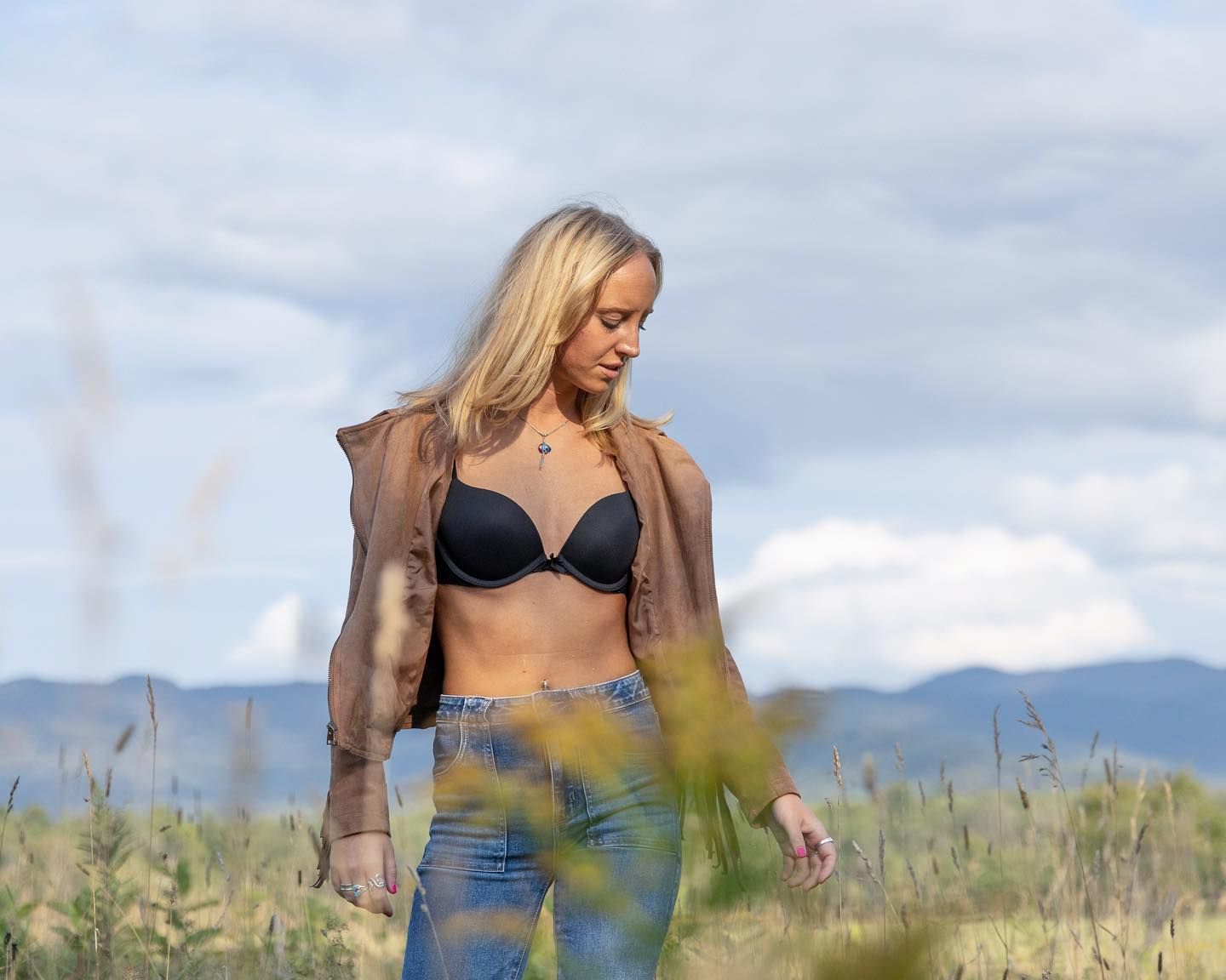 Person in casual attire walking through tall grass with mountains in the background.