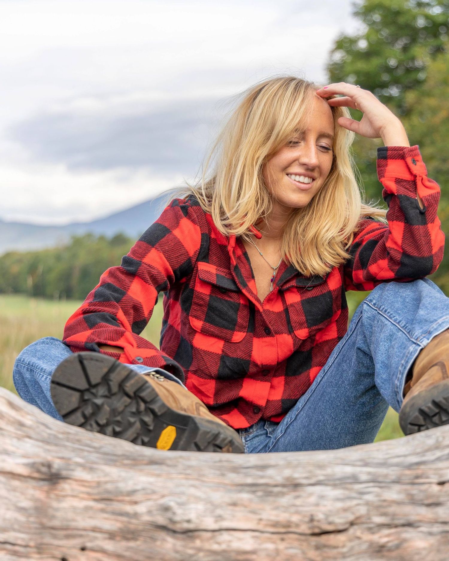 Person in red flannel shirt relaxes on fallen tree with mountains in background.