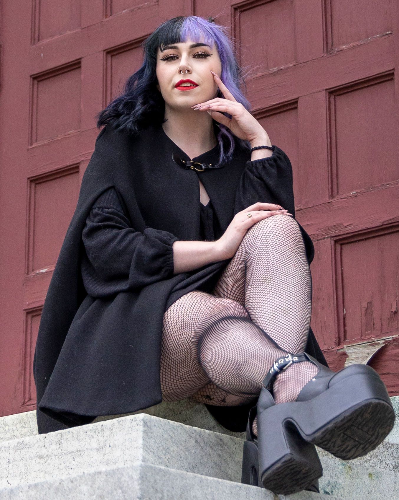 Gothic fashion pose on concrete steps wearing fishnet stockings and black cape against red door.
