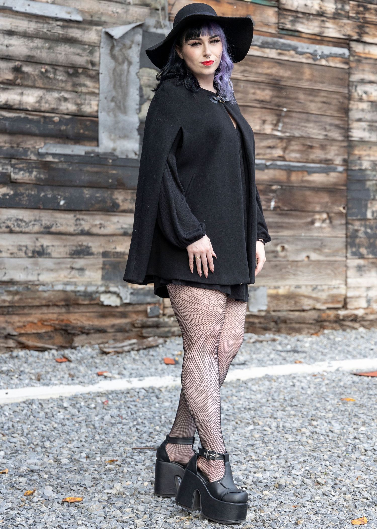 Gothic fashion model in black cape and hat poses against rustic wooden wall wearing platform shoes.