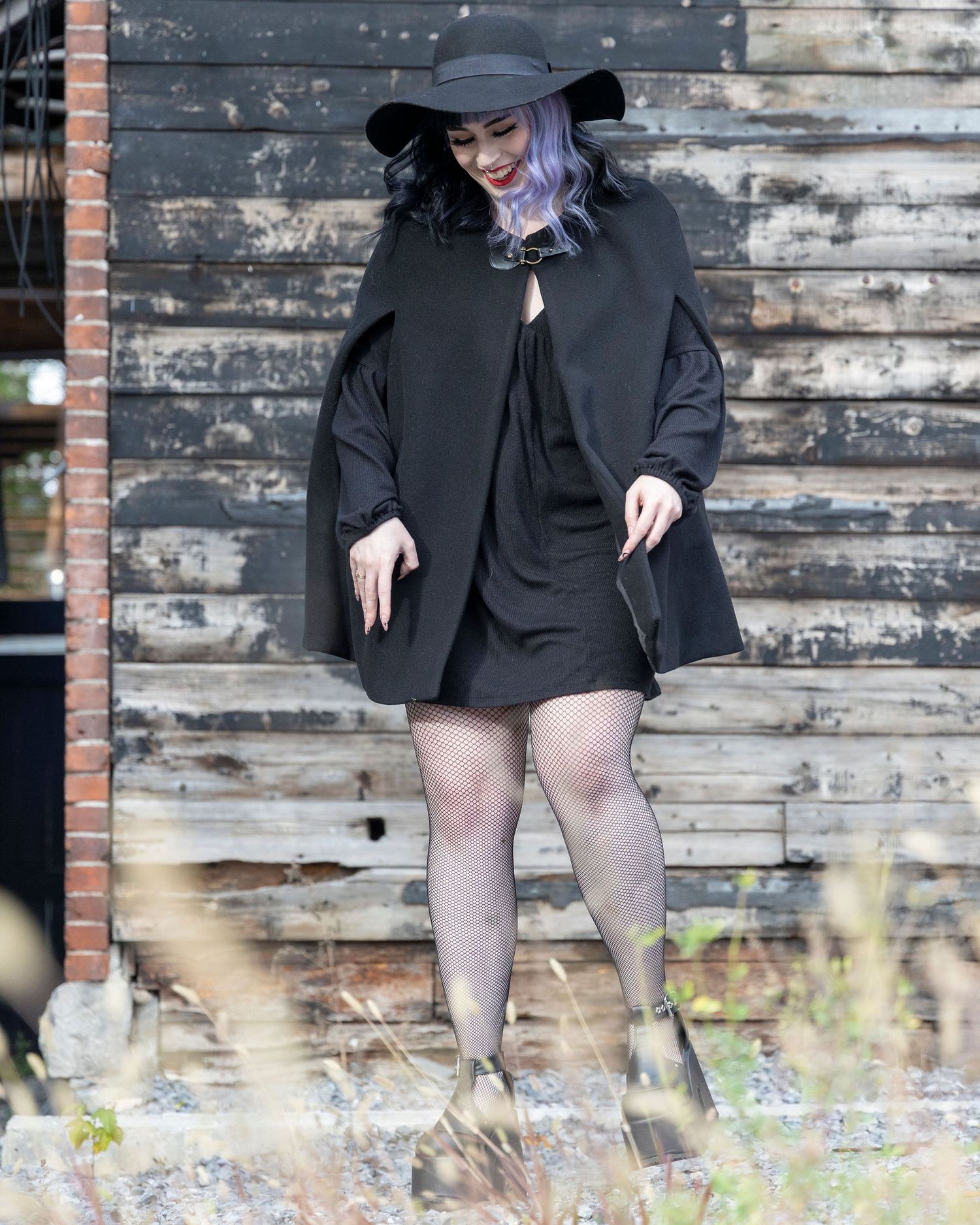 Full-length gothic style portrait in black cape and hat against weathered wood backdrop.