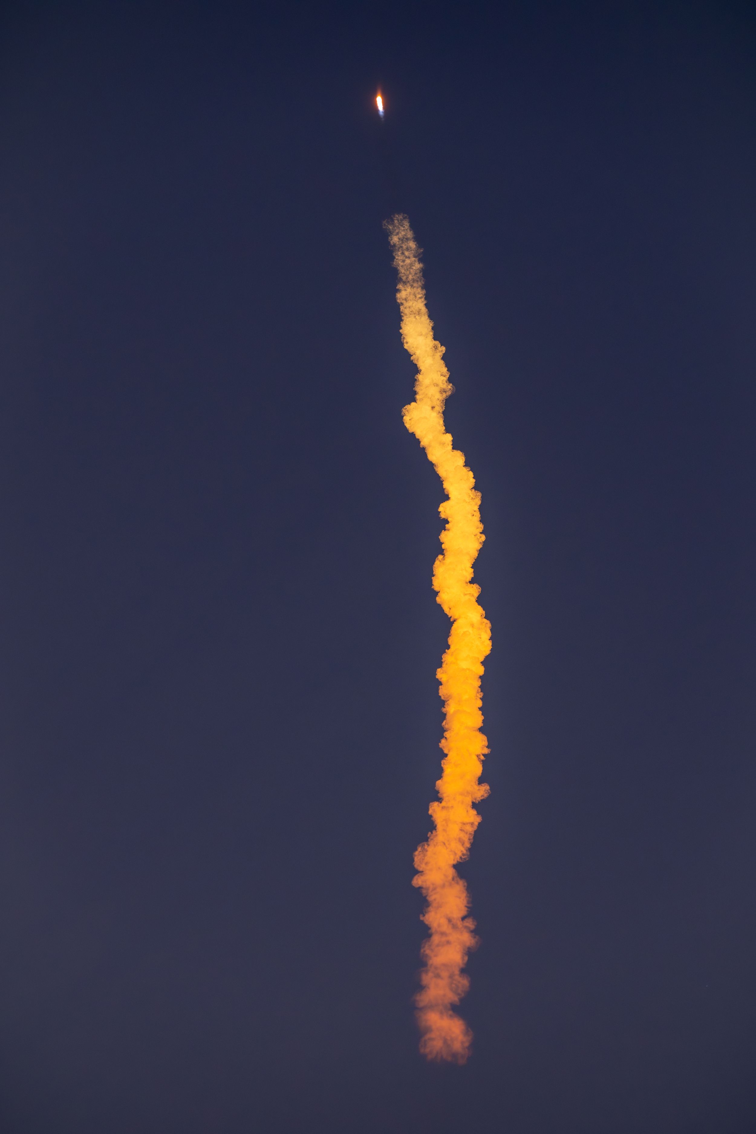 Rocket launch trail glowing orange and yellow against dark blue sky.