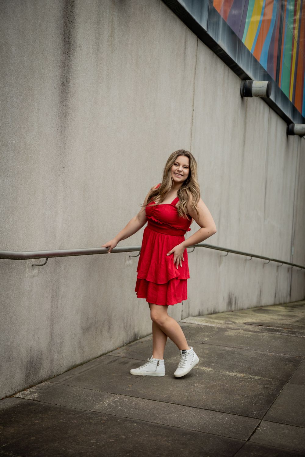 Smiling young woman in a red strapless dress leans against a railing on an urban sidewalk with colorful mural.
