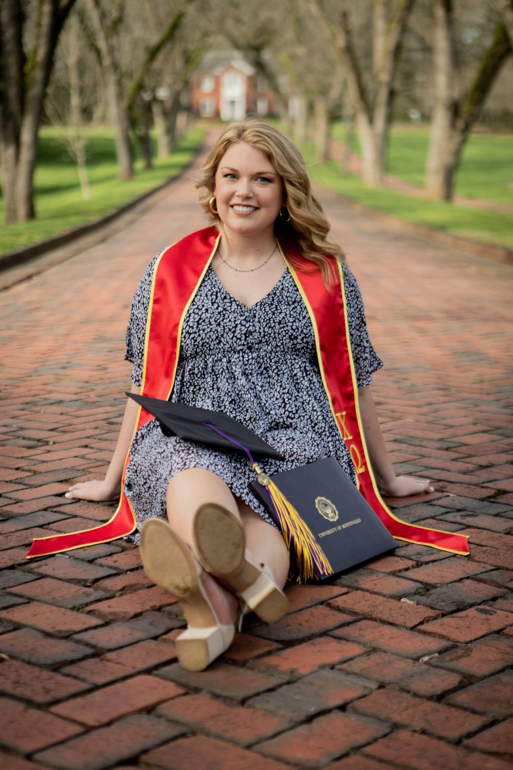 Smiling college graduate sitting on brick pathway in red graduation regalia holding diploma and cap.