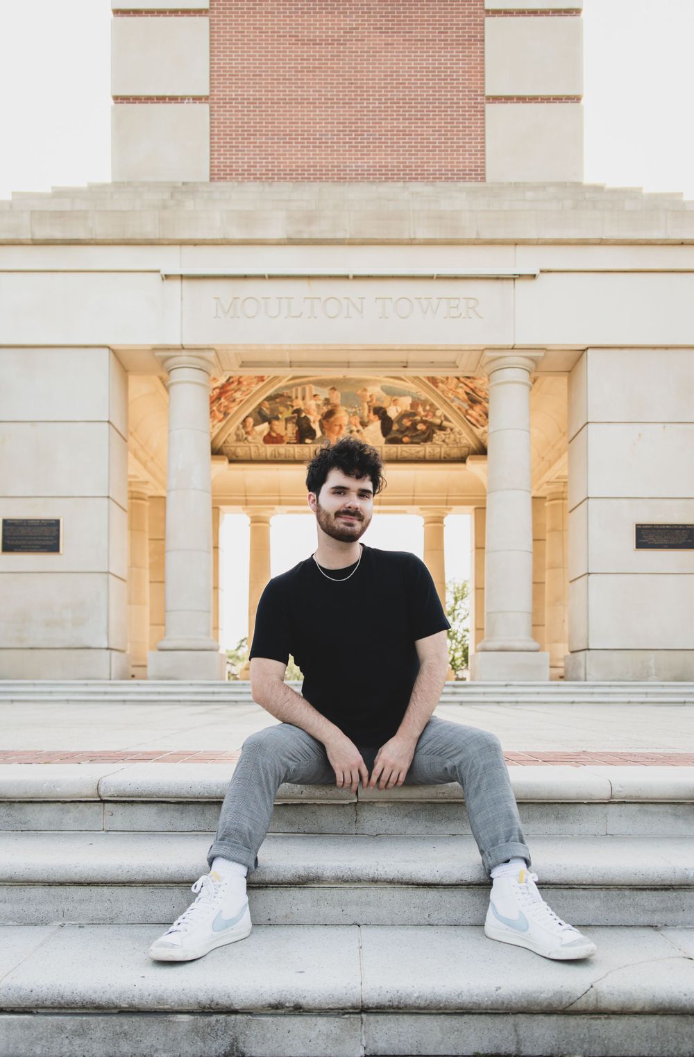 Young man in black t-shirt and jeans sits on steps in front of Moulton Tower for graduation photos.