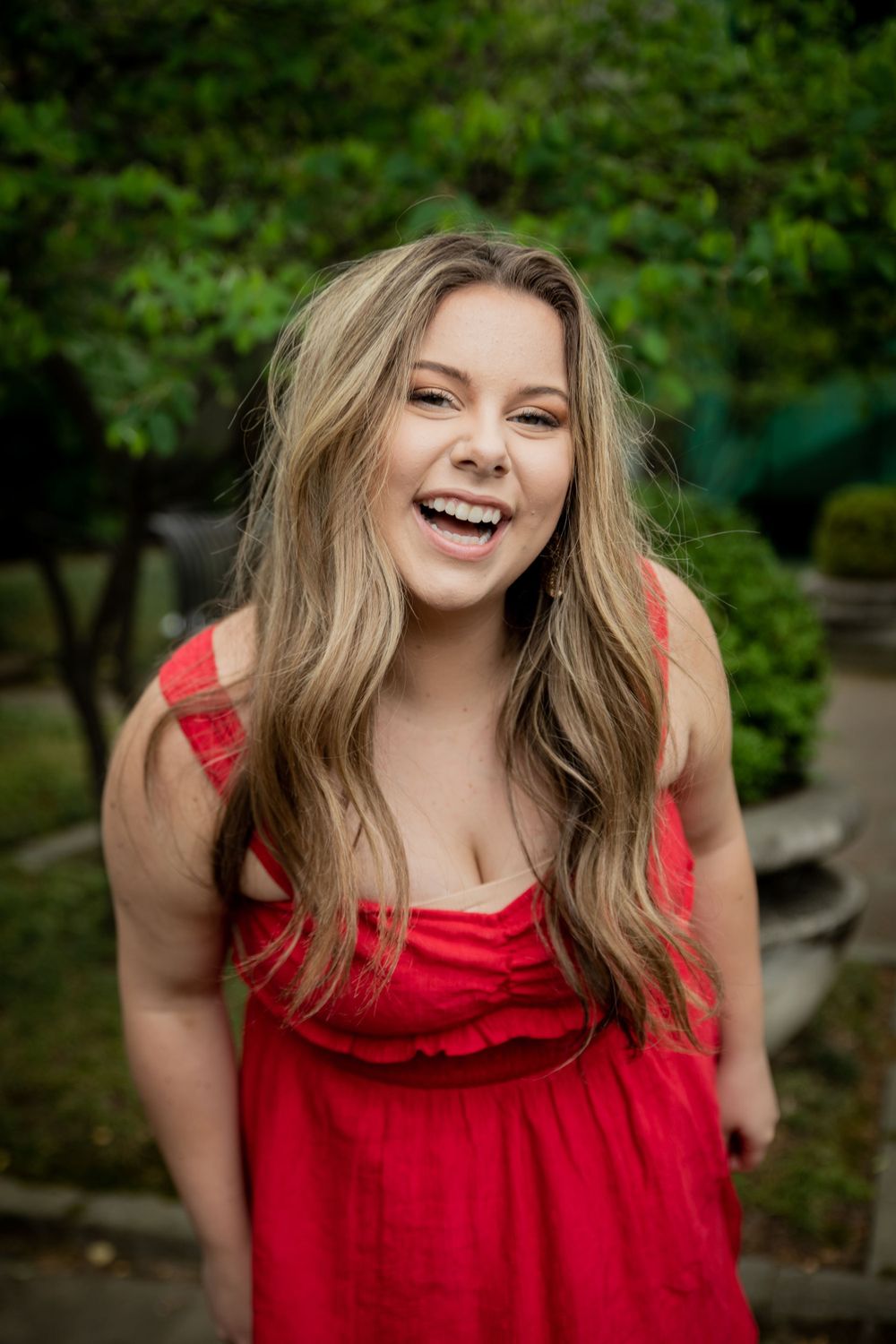 Laughing young woman with long blonde hair wearing a red dress poses outdoors in a lush green garden.