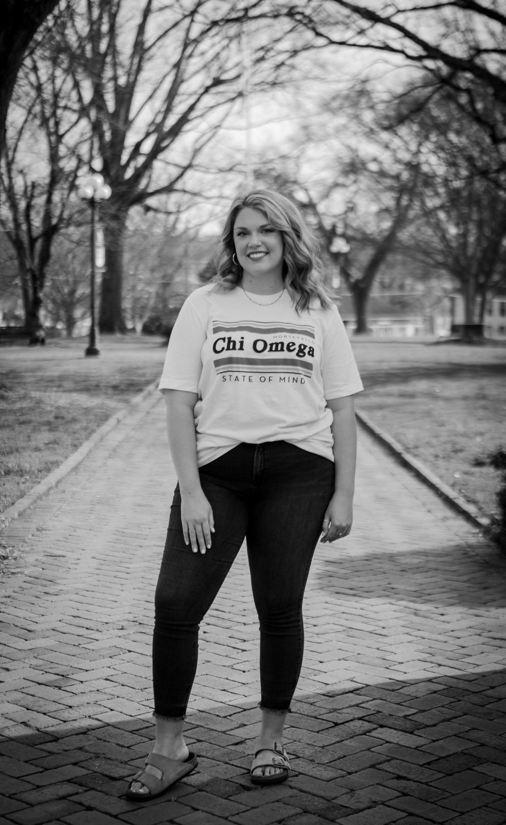 Young woman wearing Chi Omega sorority t-shirt posing on brick pathway in black and white photo.