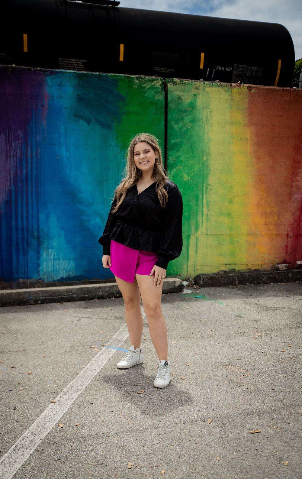 Young woman in black top and pink shorts posing in front of vibrant rainbow painted mural outdoors.