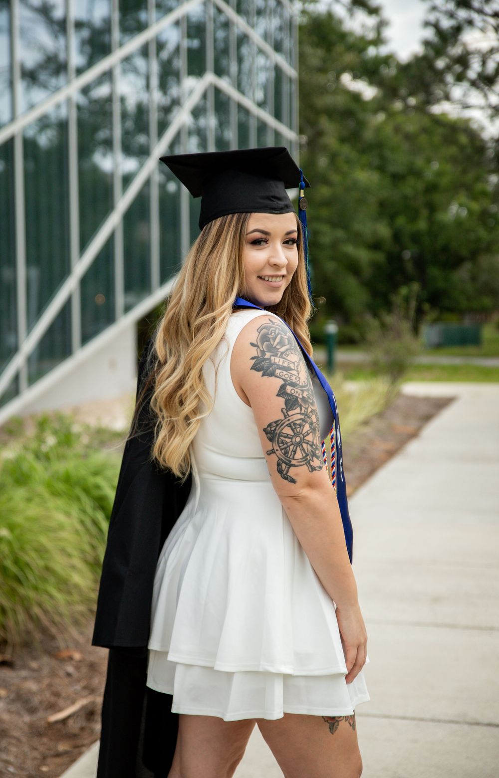 Graduate in white dress and black cap and gown shows off arm tattoo while posing outside a modern building.