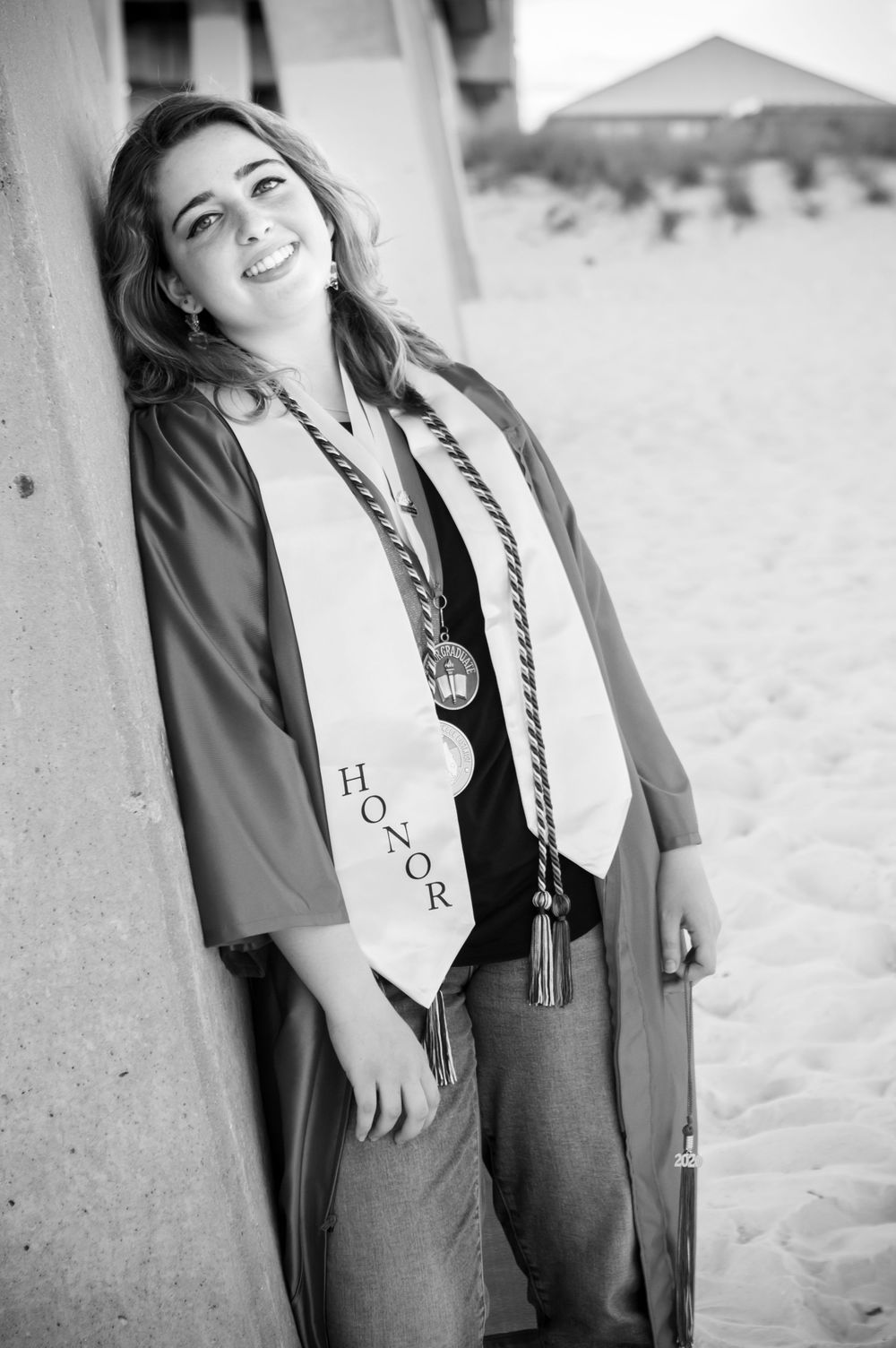 Smiling female graduate in cap and gown with honor stole leaning against wall at beach in black and white.