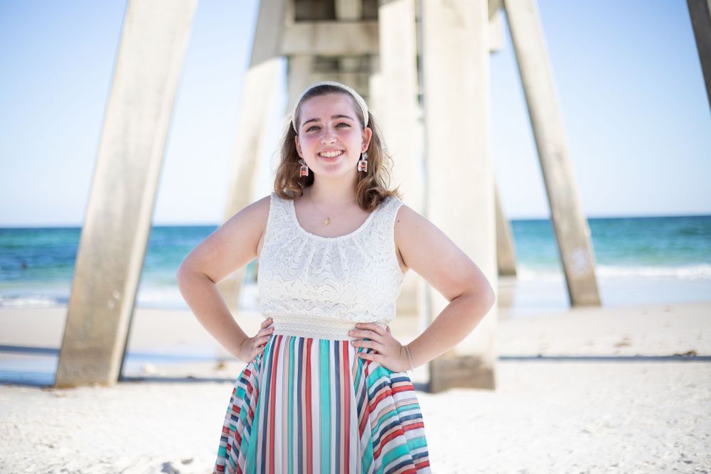 Smiling teenage girl in white lace top and colorful striped skirt posing under beach pier.