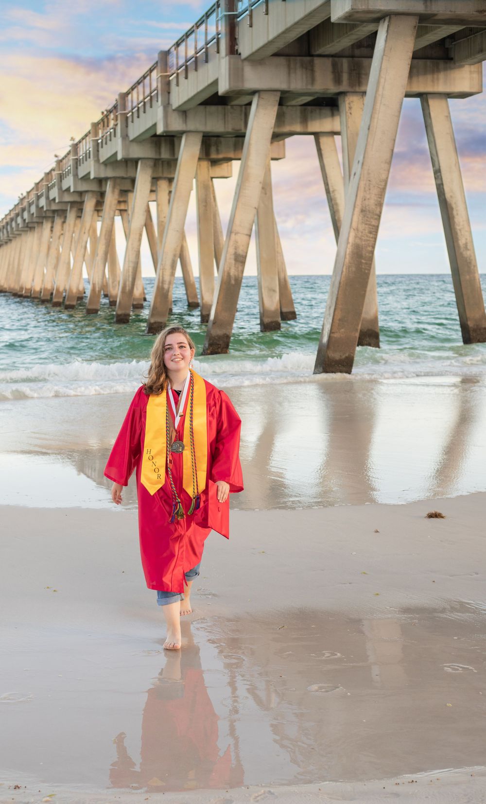 Female high school graduate in red cap and gown walking on beach near ocean pier at sunset.