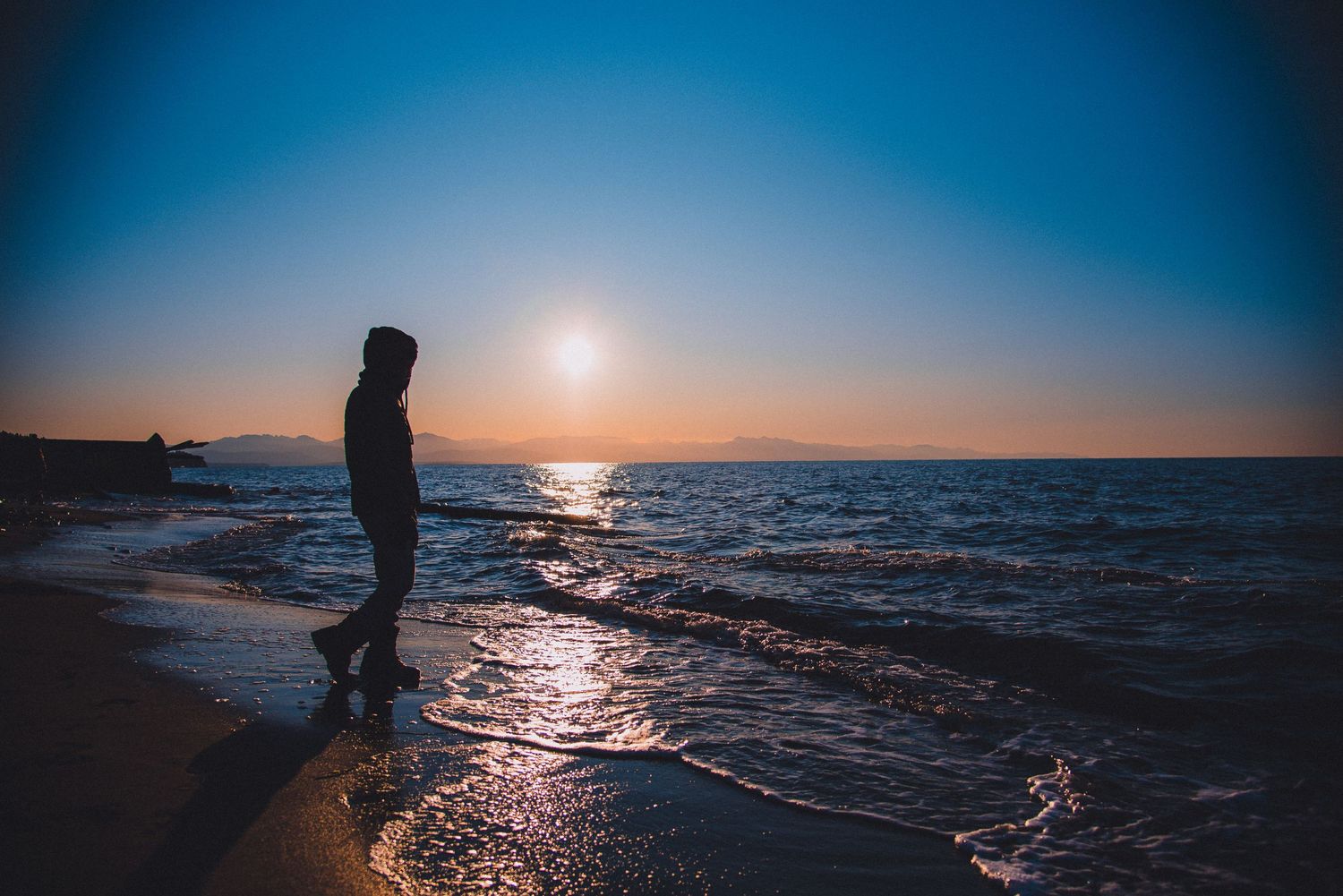 Silhouetted figure walking on a beach at sunset with a calm ocean.