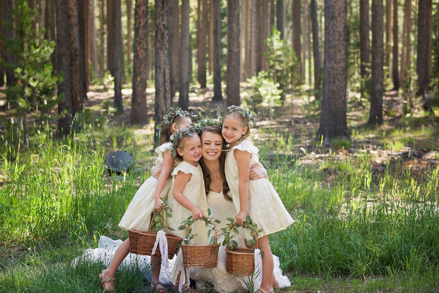 Three flowergirls in white dresses pose together in a sunny forest clearing.