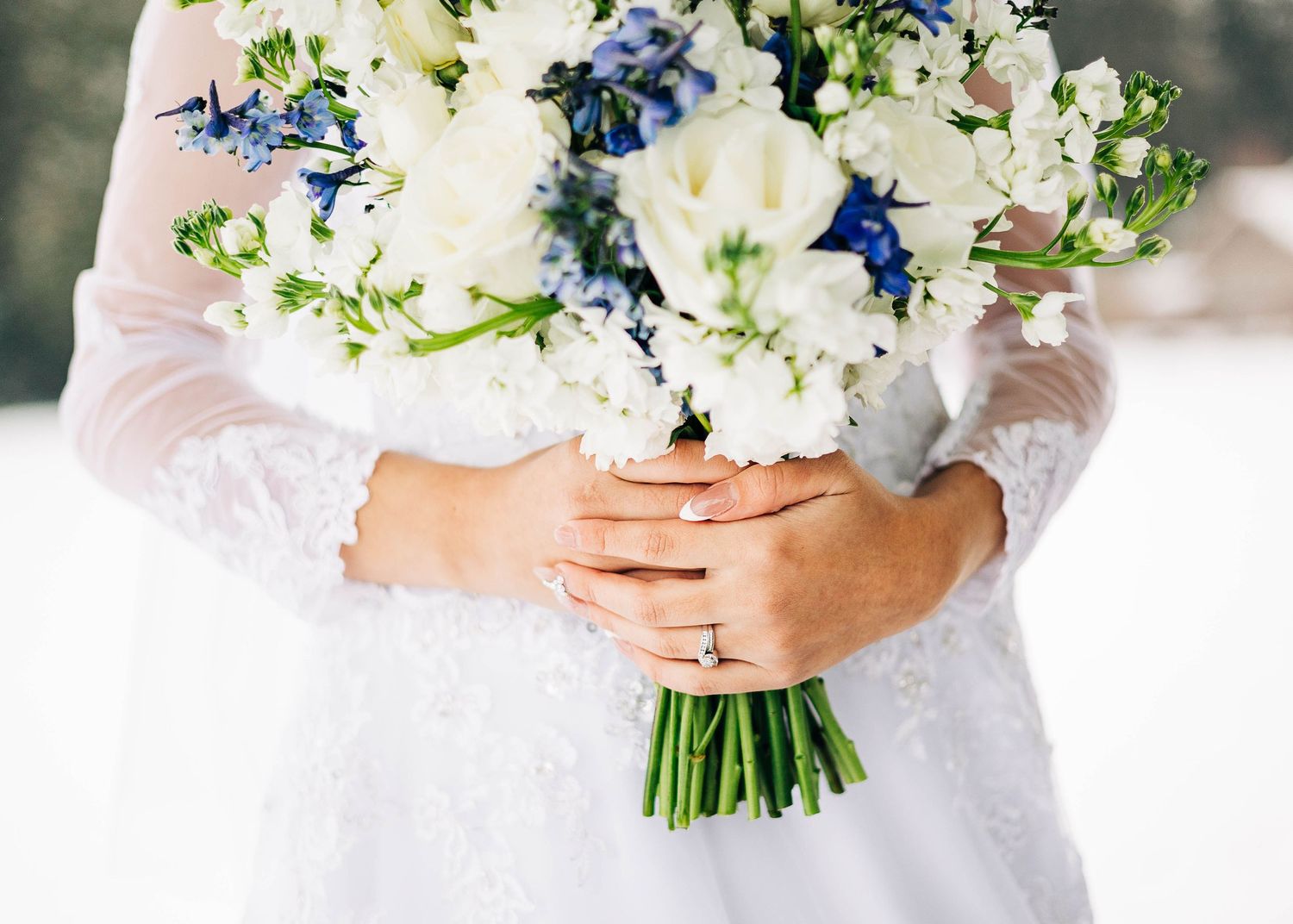 A white and blue wedding bouquet with delicate florals held by hands wearing a lace sleeve.