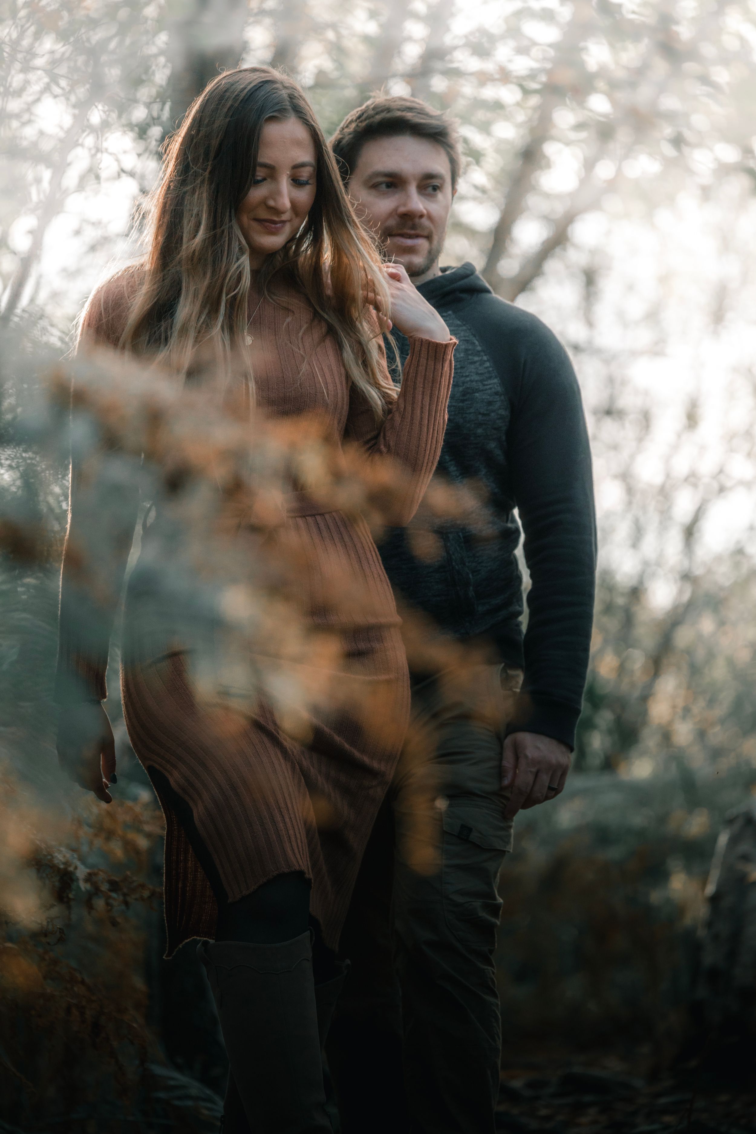 Couple standing close together in a misty autumn forest setting with warm lighting and blurred foliage.