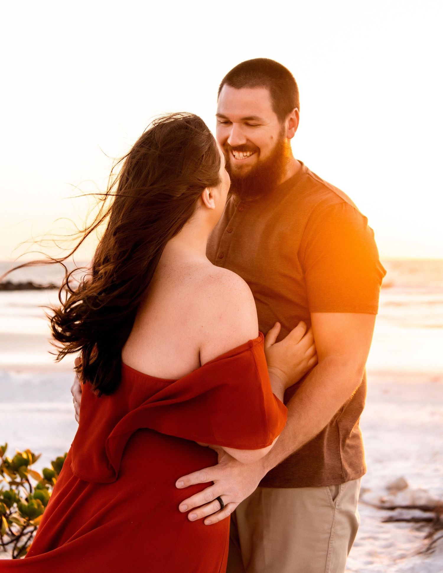 Smiling couple embracing at beach sunset, woman in off-shoulder red dress and man in casual khaki outfit.