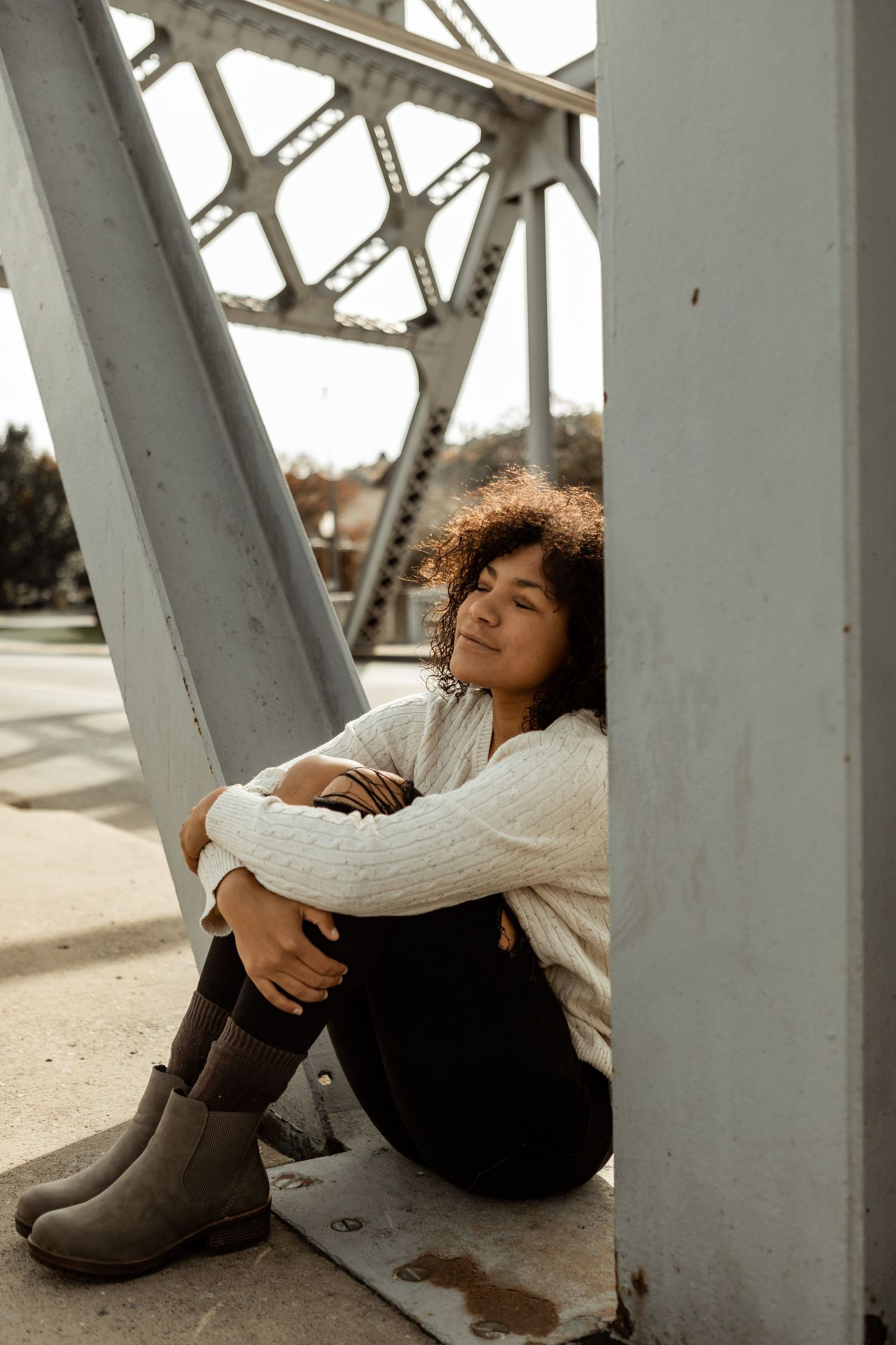 Person in white sweater and boots sitting against structural support beam outdoors.