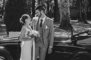 Classic black and white wedding portrait by vintage car with elegant attire and floral bouquet.