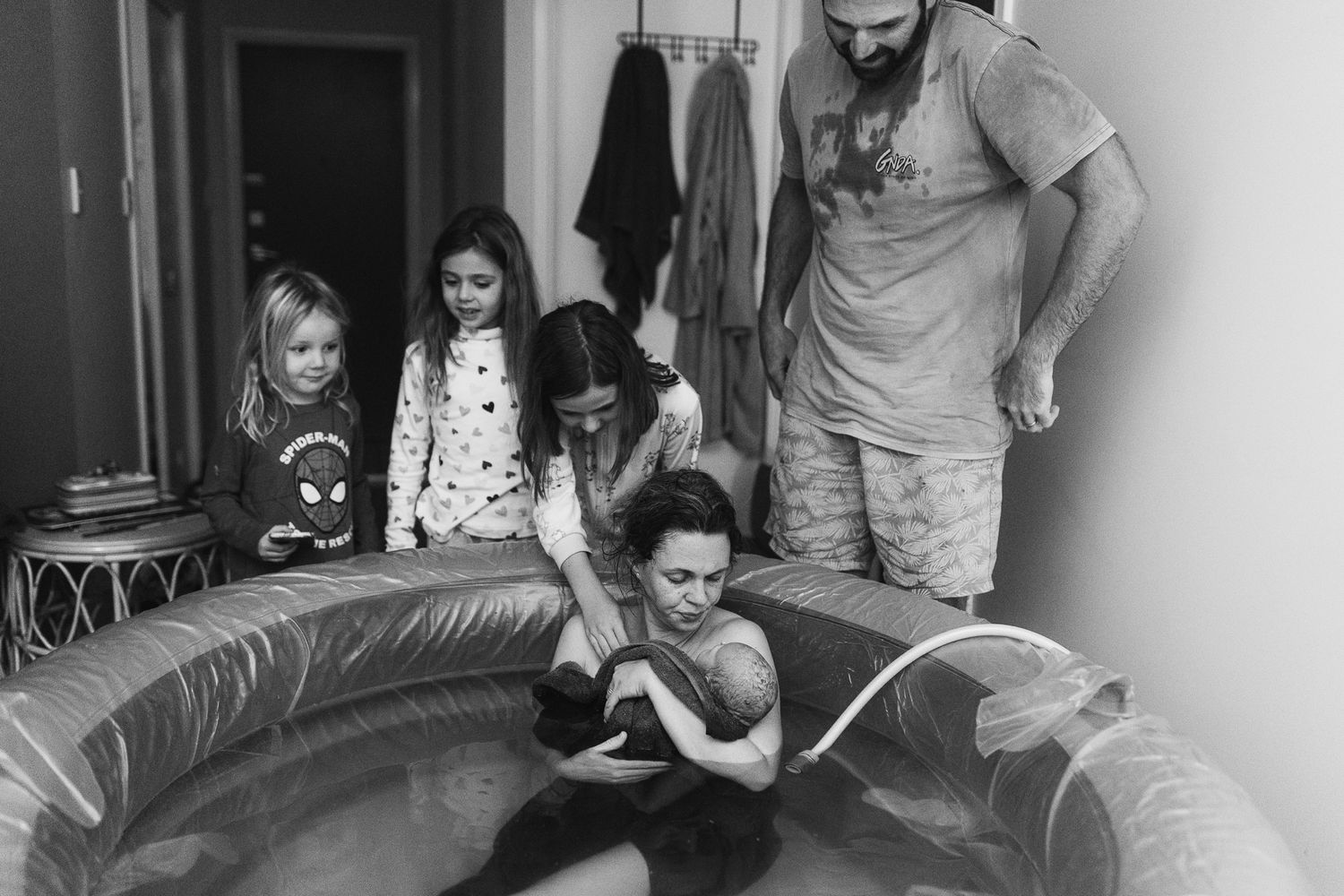 Home birth with family surrounding mother in birth pool holding newborn baby in black and white