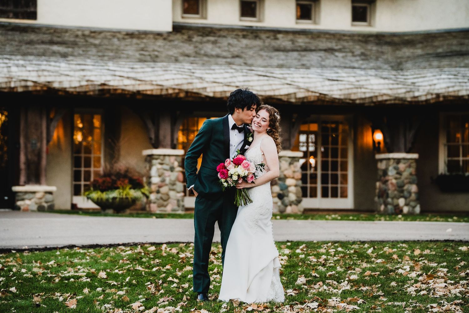 Wedding couple embracing outside stone building with red bouquet in autumn.