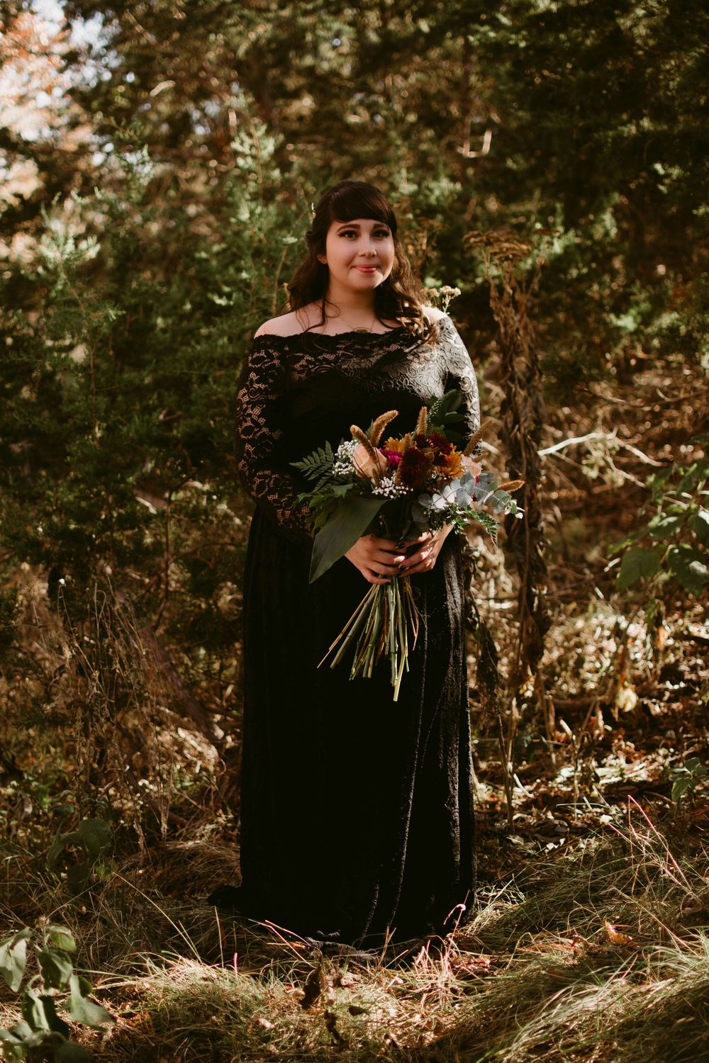 Full-length portrait in black lace dress holding autumnal bouquet in woodlands.