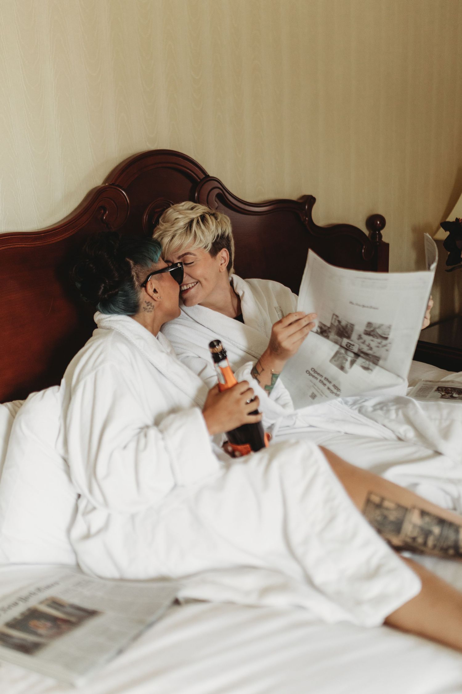 A couple in white bathrobes relaxes in bed while looking at newspapers and sharing a drink.