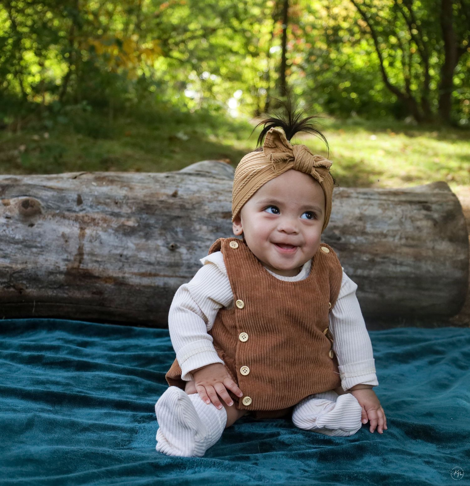 Baby in brown vest sitting on blue blanket outdoors with sunlit forest background.