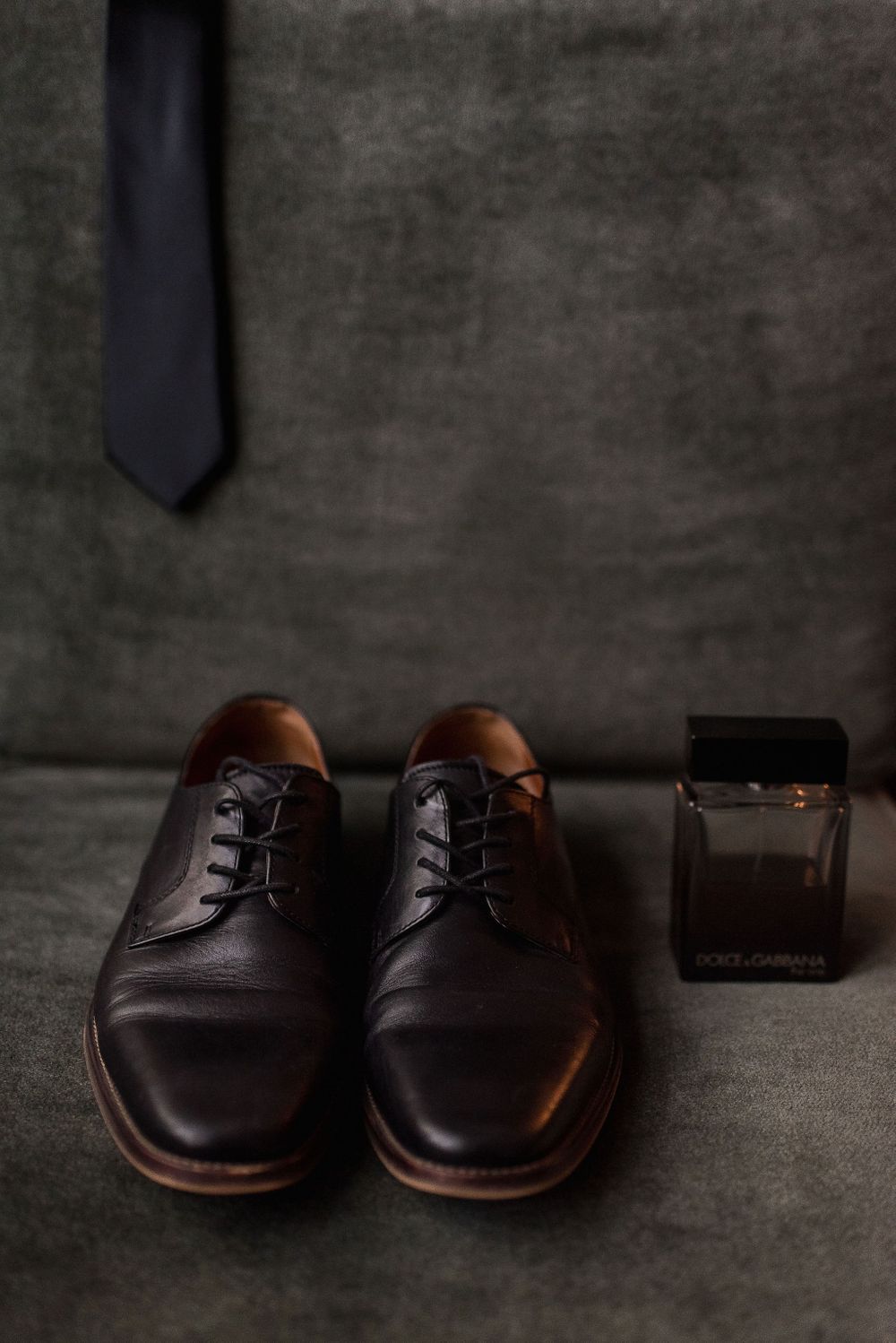Groom accessories including black dress shoes, tie and cologne on dark background.